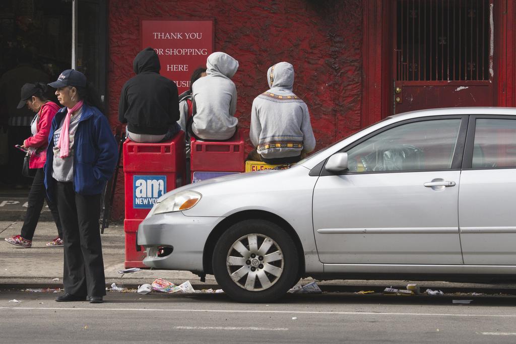 Three hooded young people sit with their backs towards me ontop of newspaper dispensing boxes. A silver car in the foreground with many items of litter on the street. A sign in the background says “Thank you for shopping here”.