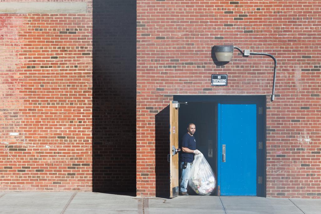 A man carries a bag of trash through a door to the street. A wide view of a red brick building, with “No trespassing” sign over the door.