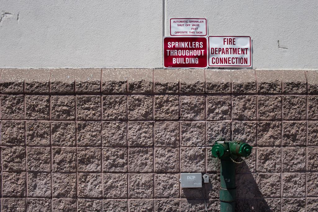 A wall with various light and geige textures, with bright red signs reading “SPRINKLERS THROUGHOUT THE BUILDING” and “FIRE DEPARTMENT CONNECTION”.