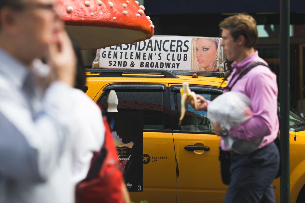 Two men walking in opposite directions, one holding his chin, the other a half eaten banana infront of a “Gentlemen's club” advert—sitting provocatively on top of a taxi in the background. A giant red and white speckled mushroom is near the foreground.