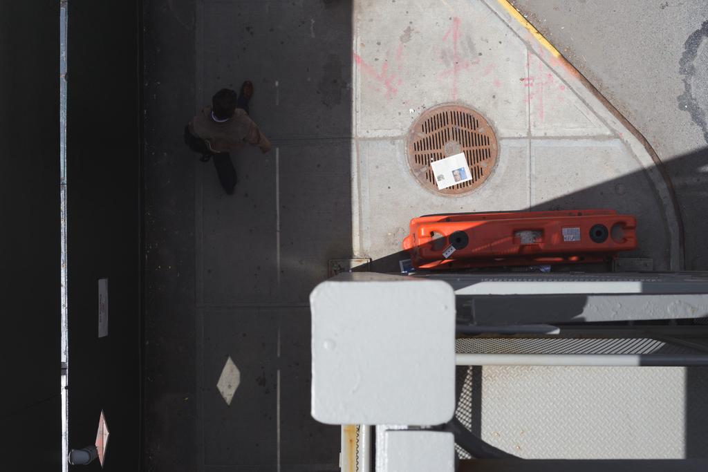 A top down view of a man walking on the sidewalk in Chelsea. An orange road barrier and newspaper lies over a circular drain cover.