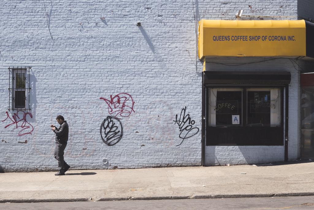 A white brick wall with man walking. A yellow sign reads “Queens coffee shop of Corona inc”.