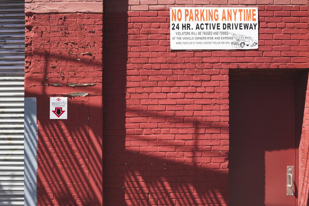 A red painted wall with intricate light and shadow and sign reading “NO PARKING ANYTIME”.