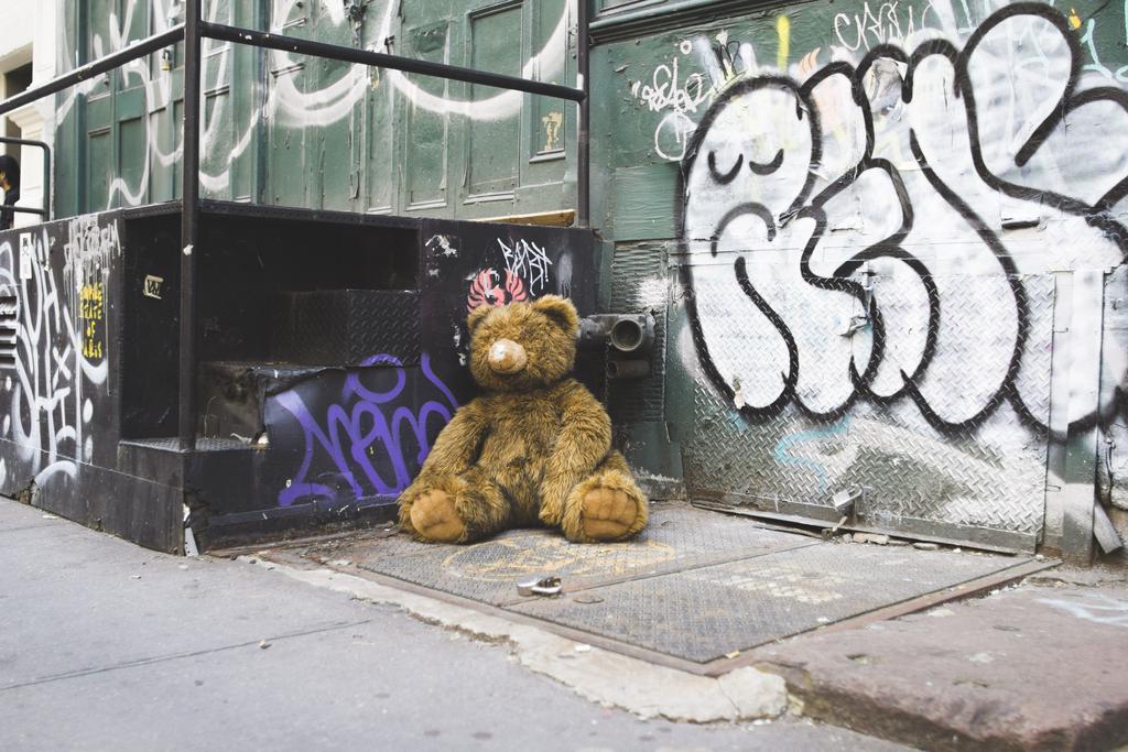 A giant teddy bear lying in the corner of a street, arguably looking sad, with a sad face graffitied on the wall behind.