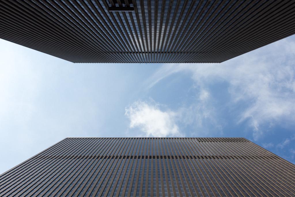 Looking up between two tall buildings in Midtown. A perfectly symmetrical view of concrete and glass, with a blue, cloudy sky in the middle.