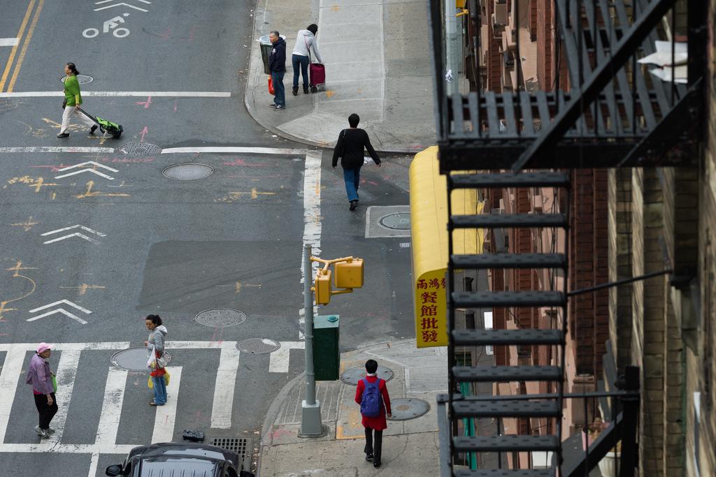 An overhead view of people walking along the sidewalk and street in different directions. A fire escape is in the foreground.