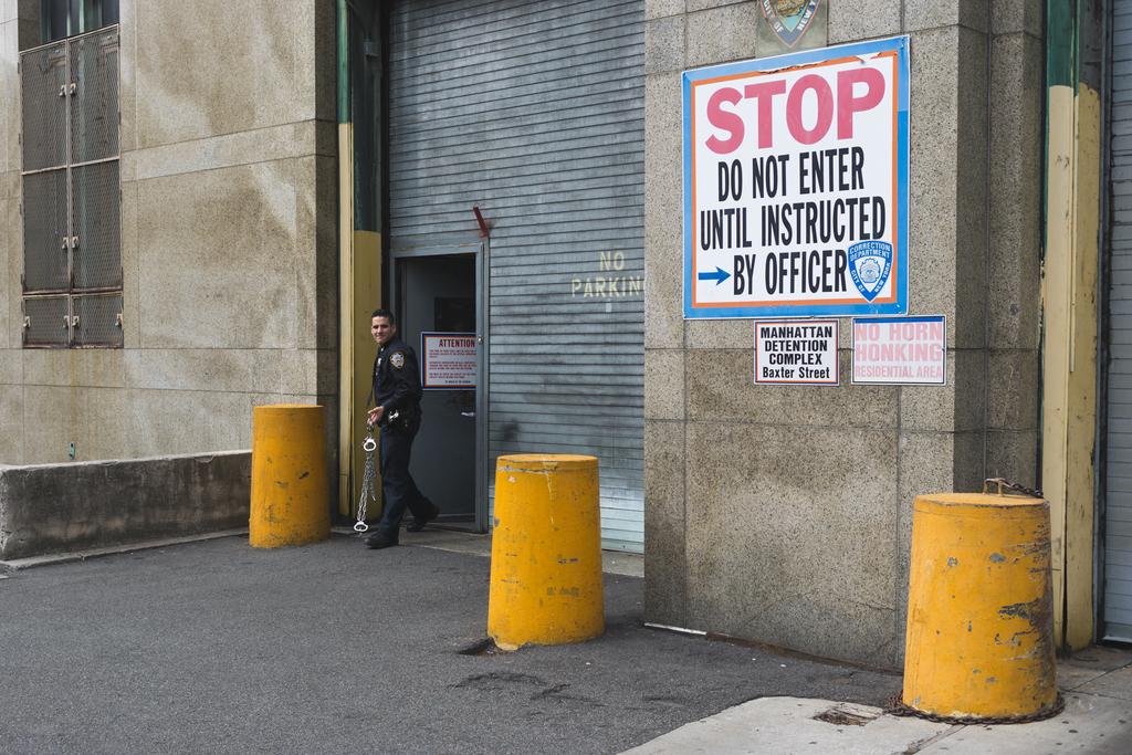 A policeman with a wry grin exits the Manhattan detention complex holding a chain of handcuffs, swingingly freely in motion. A sign on the building reads “STOP. DO NOT ENTER UNTIL INSTRUCTED BY OFFICER”